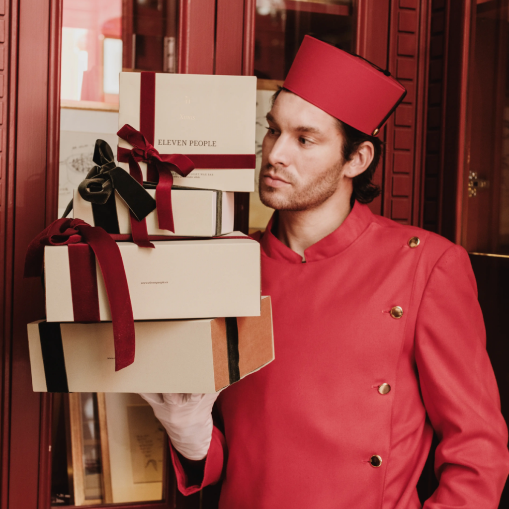 Hotelportier in roter Uniform trägt mehrere elegant verpackte Geschenkboxen von Eleven People mit Schleifen – luxuriöse Geschenkverpackung und Premium-Präsentation.