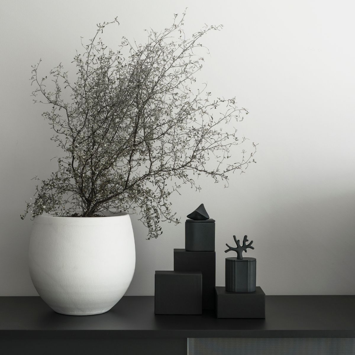 Objets décoratifs Kyuka Design – sculptures noires modernes sur socles à côté d'un grand vase blanc contenant des brindilles séchées, intérieur minimaliste.