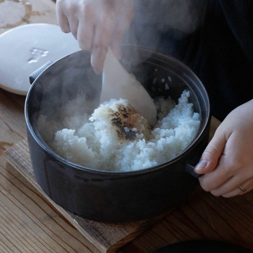 Japacolle Donabe – schwarzer japanischer Keramik-Reiskochtopf mit dampfendem Reis, traditionell serviert und mit einem Reislöffel gelockert.
