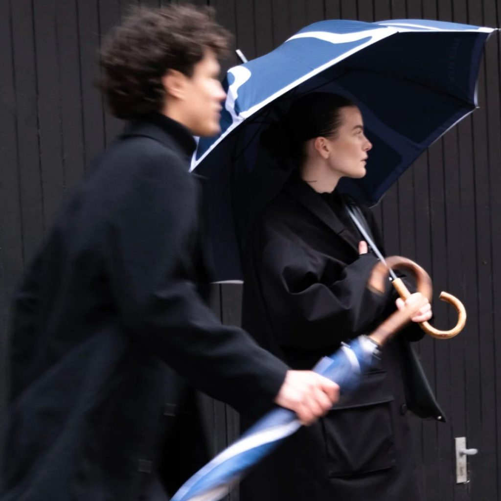 Zwei Personen in dunkler Kleidung gehen im Regen, eine Frau hält einen blauen Sapor Design Regenschirm mit Holzgriff.