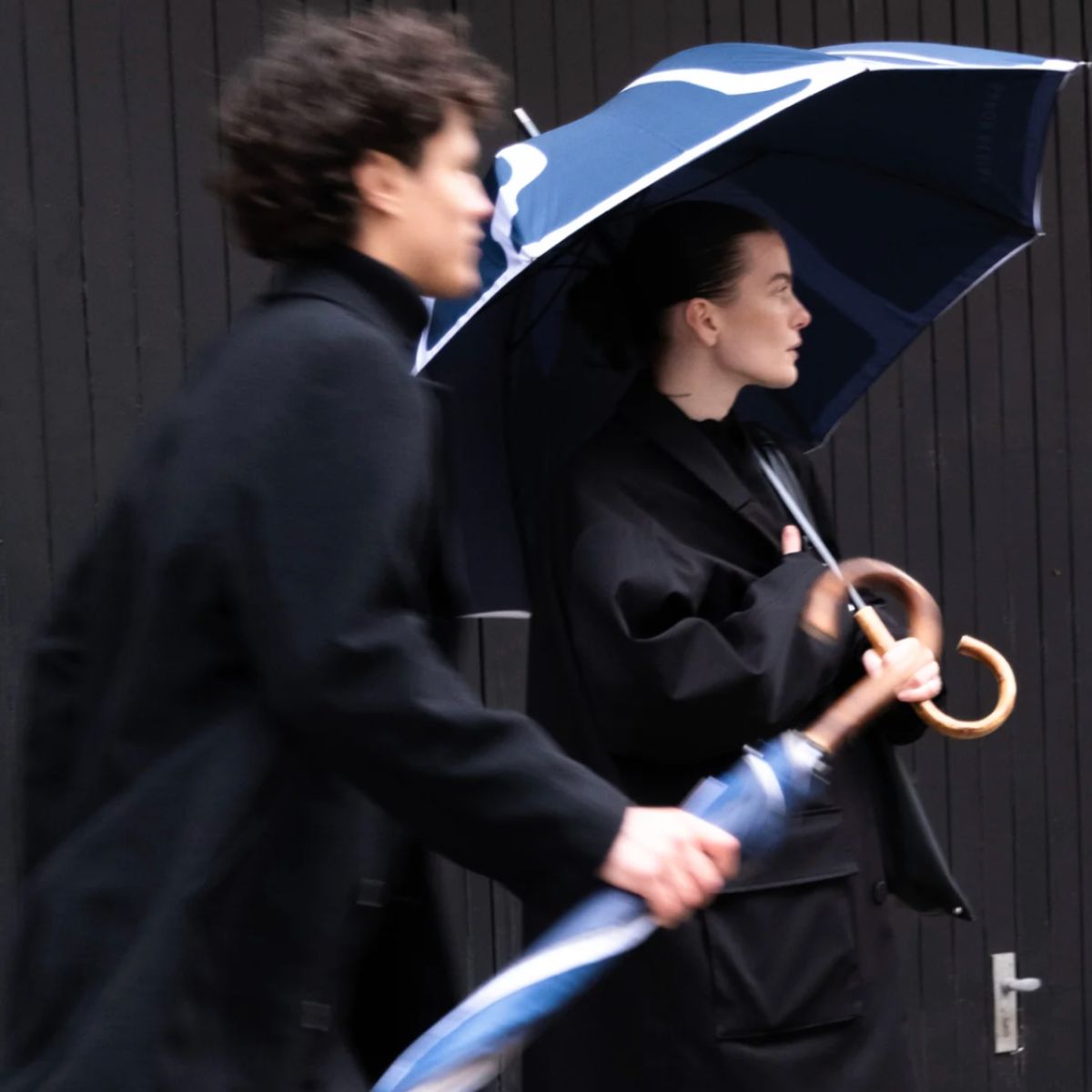Zwei Personen in dunkler Kleidung gehen im Regen, eine Frau hält einen blauen Sapor Design Regenschirm mit Holzgriff.