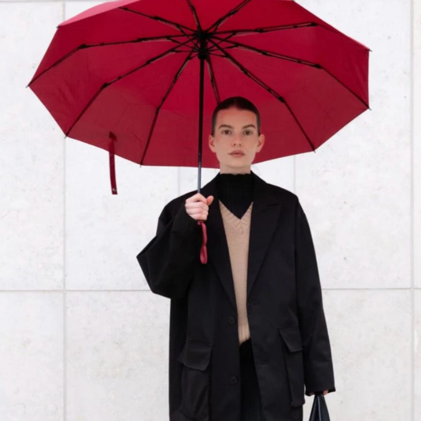 Person hält einen geöffneten roten Taschenschirm in zeitlosem Design von Sapor Design in rot und trägt einen schwarzen Oversize-Mantel vor hellem Hintergrund.