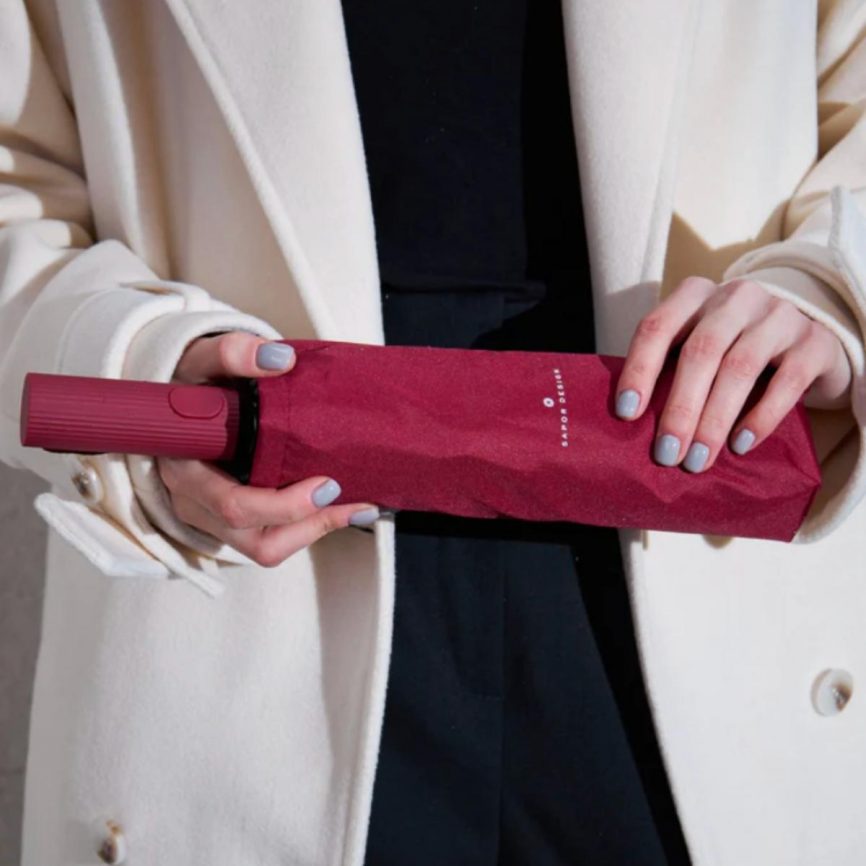 Person hält einen roten Taschenschirm in zeitlosem Design von Sapor Design in rot in der Schutzhülle, getragen zu hellem Mantel und schwarzer Kleidung.