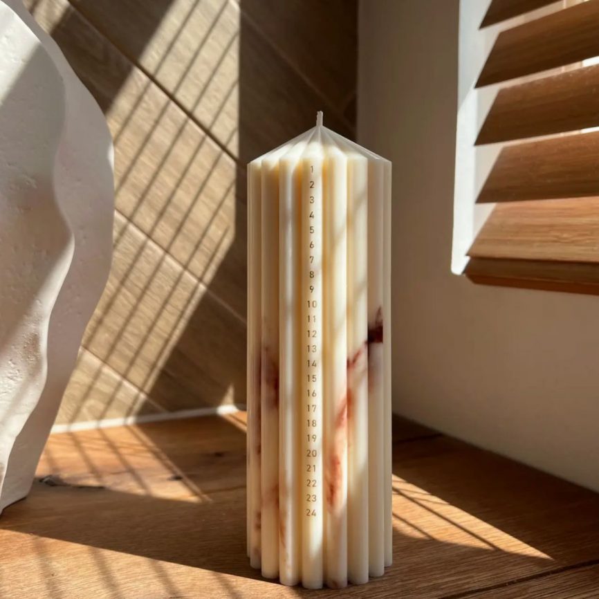 Kalenderkerze von Studio M Candles aus cremefarbenem Wachs mit einer Datums­skala von 1–24, wunderschön im warmen Sonnenlicht auf einem Holztisch platziert.