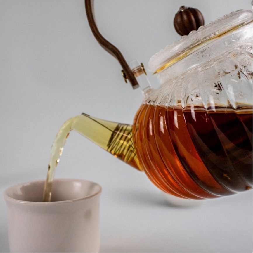 Gläserne Teekanne mit Holzgriff gießt frisch aufgebrühten Tee in eine minimalistische weiße Tasse – elegantes Teeritual mit hochwertigem Teeservice.