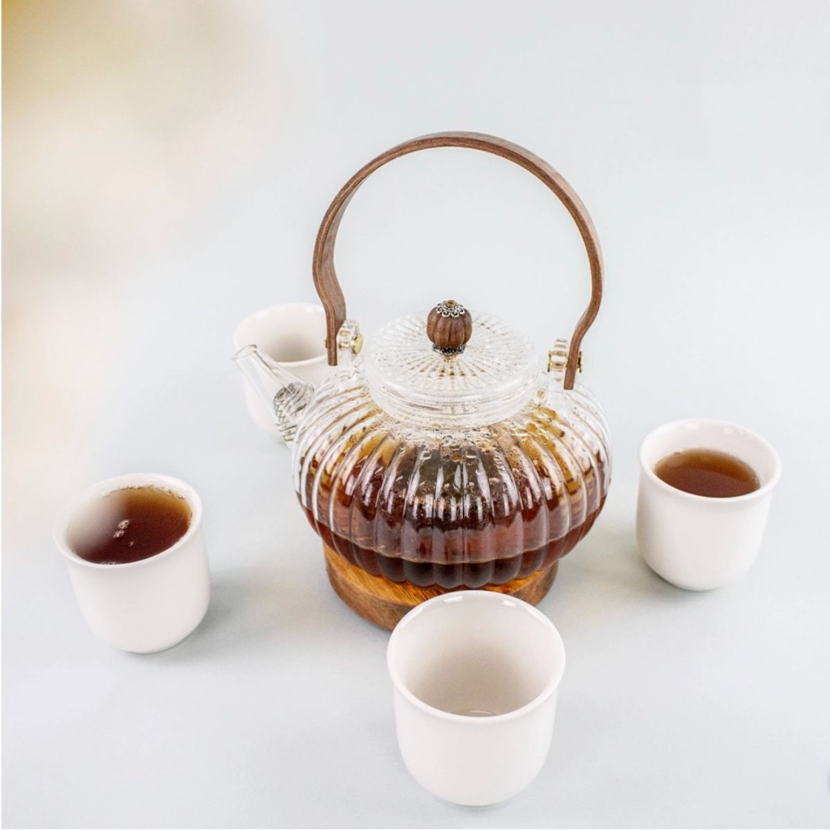 Tetera de cristal con mango de madera y cuatro tazas de té blancas sobre un fondo claro: un elegante servicio de té para ceremonias del té con estilo.