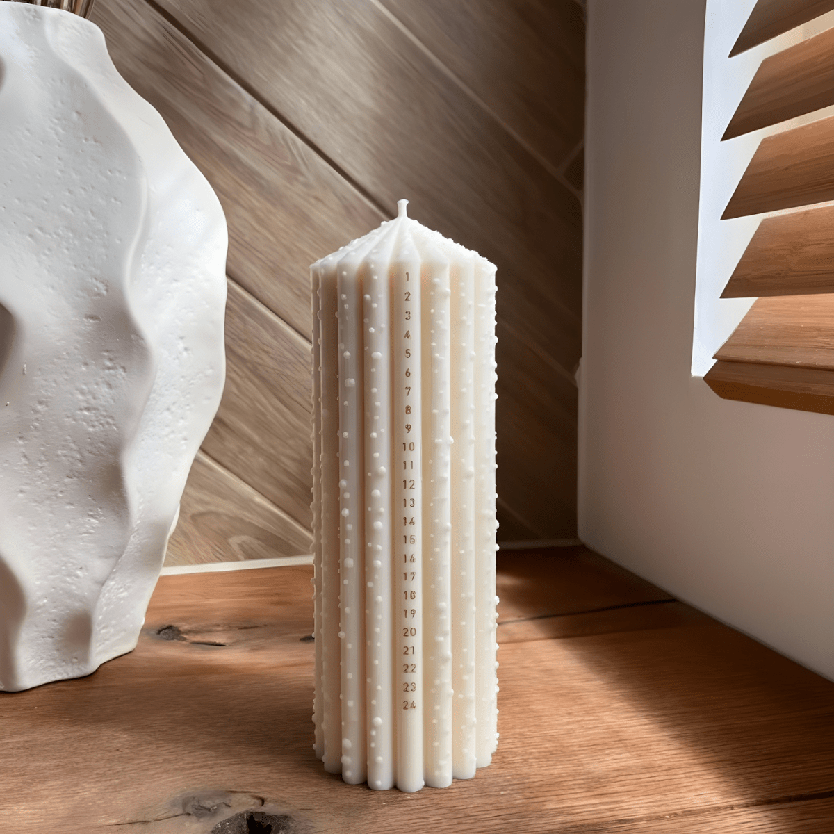 Dänische Kalenderkerze in Creme von Studio M Candles mit 1–24 Datumsskala und strukturierter Oberfläche, stehend auf einem Holztisch im warmen Tageslicht.