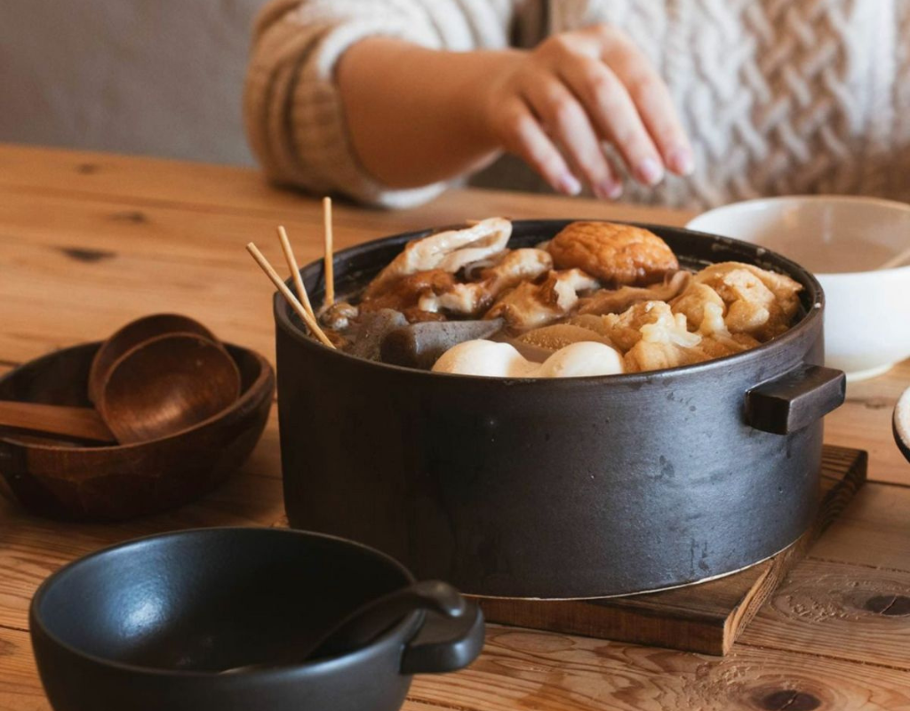 Traditioneller schwarzer Kochtopf mit Oden und verschiedenen Zutaten auf Holztisch, umgeben von Keramikschalen und Holzlöffeln – gemütliche japanische Essensszene.